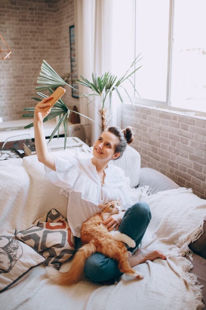 From above of smiling female in casual clothes sitting on bed and stroking adorable cat while taking selfie in cozy bedroom at home