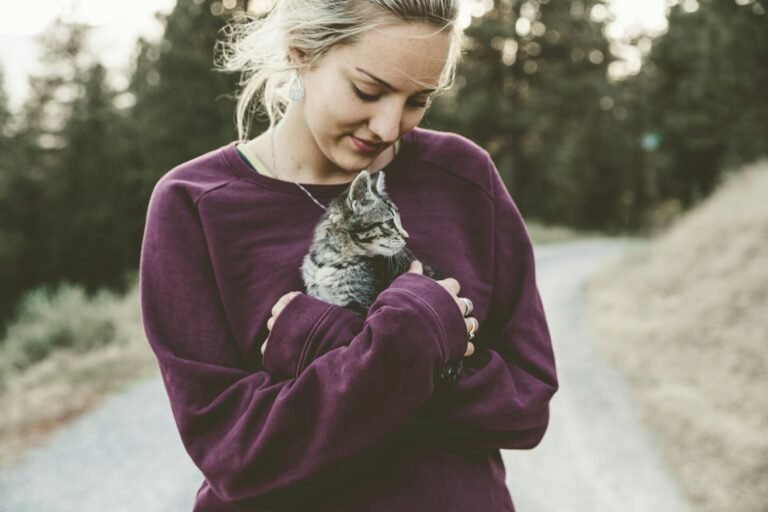 Young woman in a purple sweater smiling down at a kitten she's holding outdoors on a pathway.